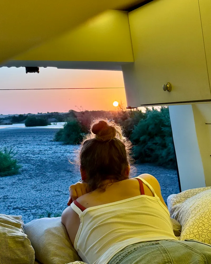 Voyageuse admirant le coucher du soleil depuis le lit du van aménagé, moment détente en vanlife au Maroc.